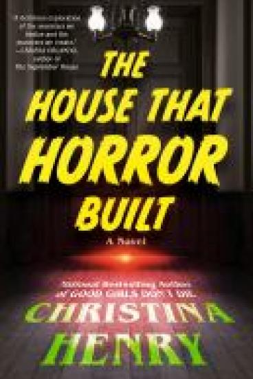 Book cover for The House that Horror Built, featuring a realistic illustration of a hallway with a closed door at its end and red light seeping out from the crack beneath.  A vintage iron chandelier hangs from the ceiling before the door, and the walls feature wood panel wainscotting.  The red light from beneath the door casts an eerie glow on the floorboards of the hallway.