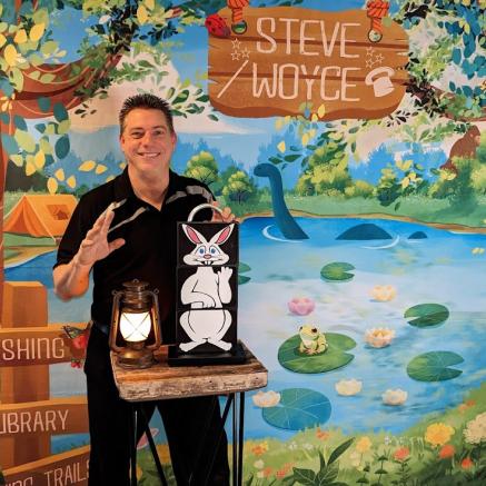 Magician Steve Woyce stands in front of a forest background, with a lantern and a picture of a white rabbit in front of him