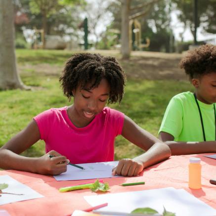 Two children coloring outside with paper on a checkered picnic table. One is wearing a green shirt and the other is a pink shirt. Grass is in the background