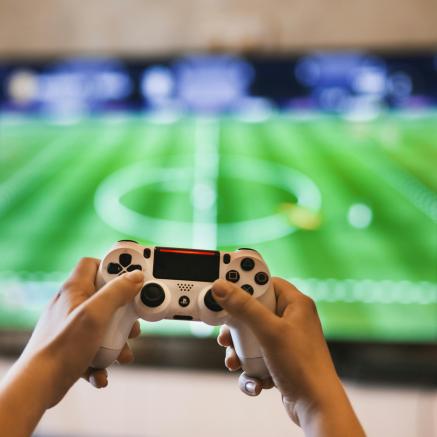 Young hands hold a Playstation Controller playing a soccer video game on the tv behind the hands