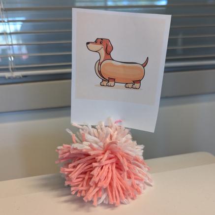 A pom-pom made of pink and white yarn is holding a cartoon picture of a dachshund.