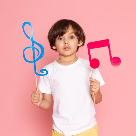 Little boy stands against a pink background holding a treble clef and two 8th notes