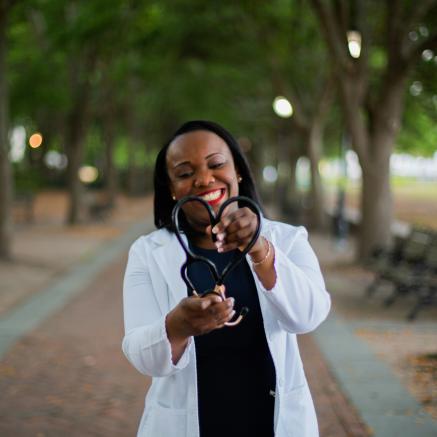 Woman in white lab coat smiles while forming stethoscope into a heart