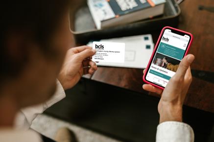 Person holding a BCLS library card and a cell phone with the BCLS Mobile website