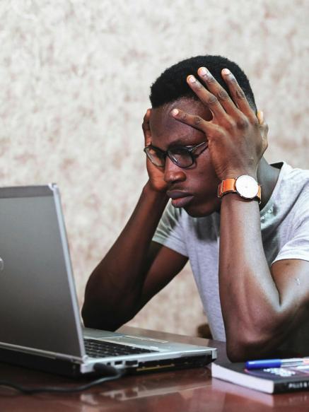 a young african-american man holds his head in frustration in front of his laptop.