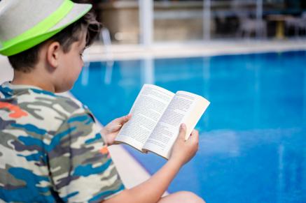 Child in blue camo bathing suit and green hat reading outside by the pool