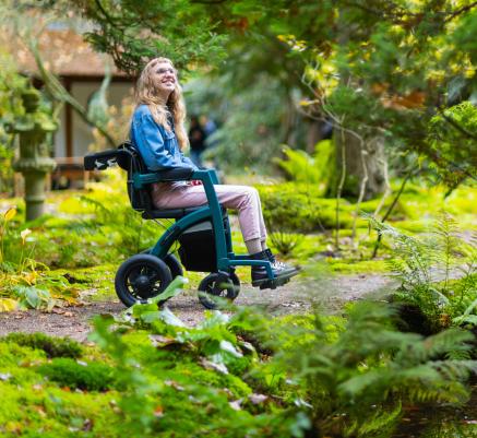 A teen girl in a wheelchair outdoors surrounded by green grass and leaves.