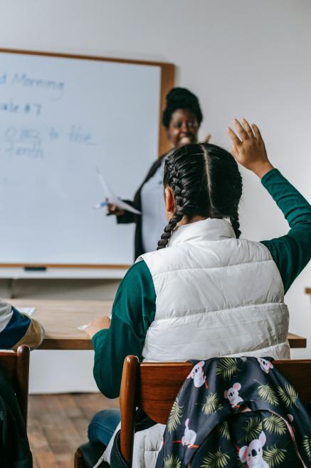 A teacher smiles at a classroom while a student raises her hand.