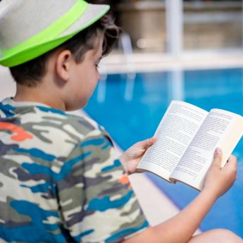 Child in blue camo bathing suit and green hat reading outside by the pool