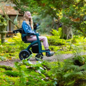 A teen girl in a wheelchair outdoors surrounded by green grass and leaves.