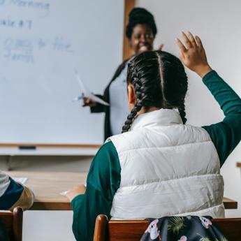 A teacher smiles at a classroom while a student raises her hand.