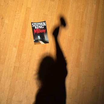 "Misery" by Stephen King on the floor with the shadow of a woman holding a mallet over her head next to it